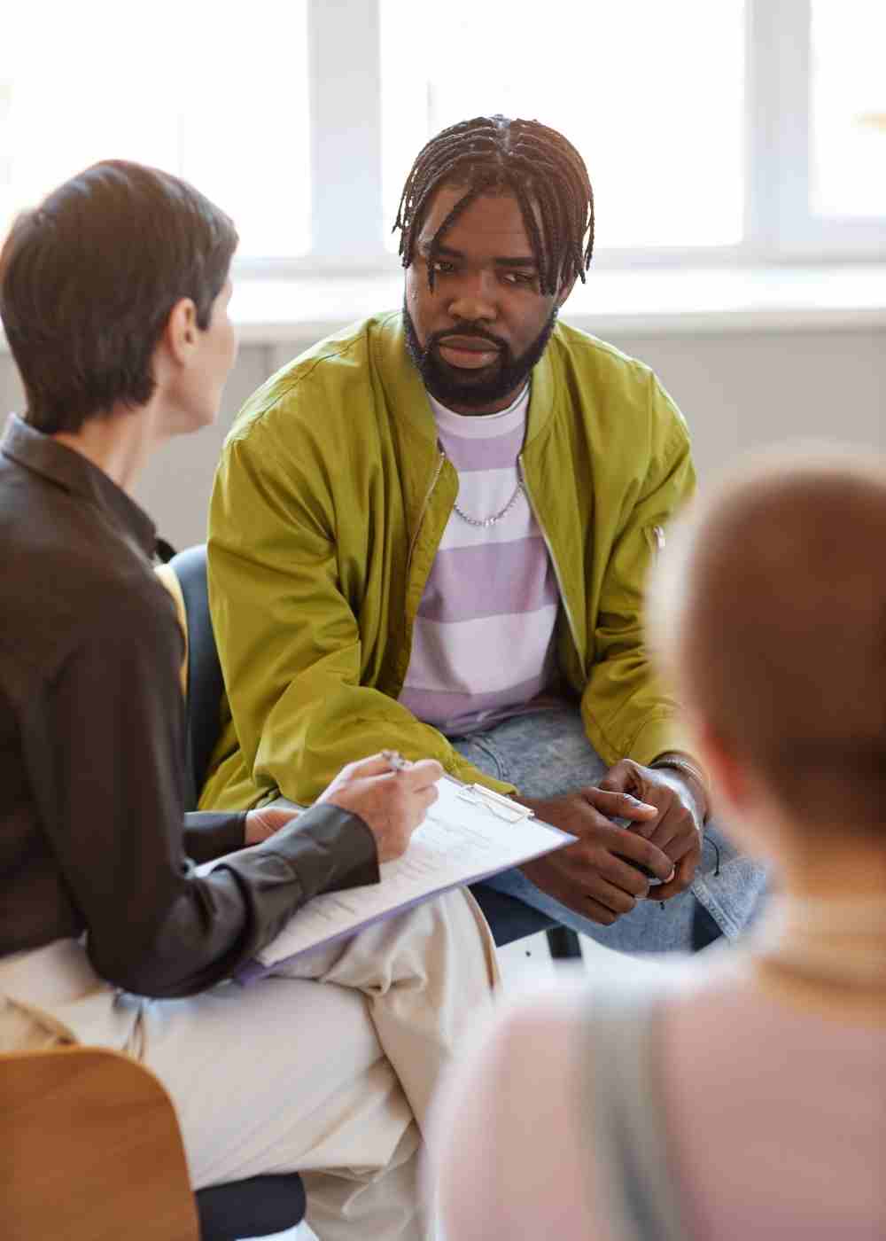 A therapist speaks with a client during an individual addiction treatment session while others participate in a support group.